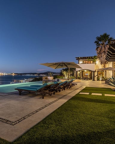 Luxury villa with illuminated terrace, infinity pool, lounge chairs, and ocean view at dusk