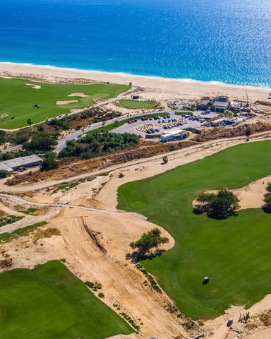 Aerial view of coastal golf course with multiple fairways, bunkers, clubhouse, parking area, and turquoise ocean beyond sandy beach.