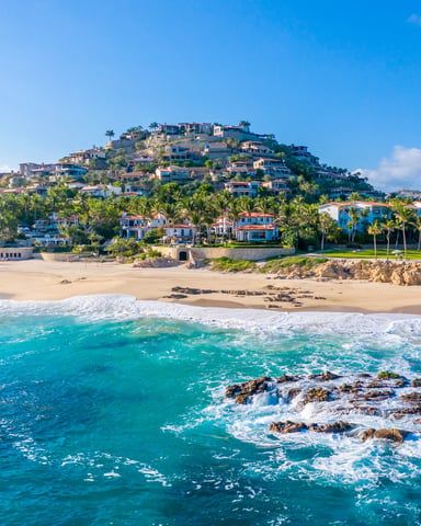 Beachfront hillside community with colorful homes, palm trees, sandy beach, and turquoise ocean waves