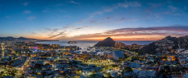 Coastal city at dusk with illuminated buildings, rocky mountains, and bay with island at sunset