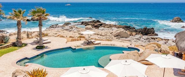 Villa de lujo con piscina infinita, tumbonas y terraza iluminada con vistas a una bahía al atardecer