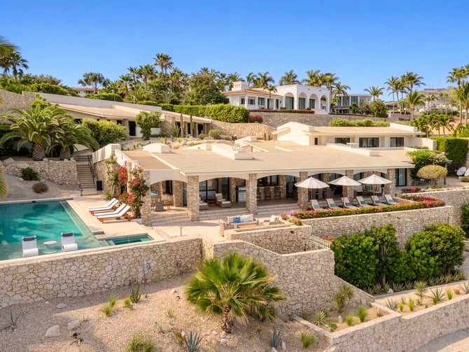 Luxury villa with stone and white architecture, swimming pool, shaded terrace, palm trees, and coastal hillside setting