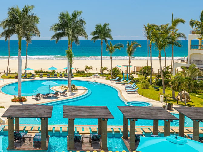 Resort pool with turquoise water, palm trees, beach umbrellas, and ocean view beyond sandy beach