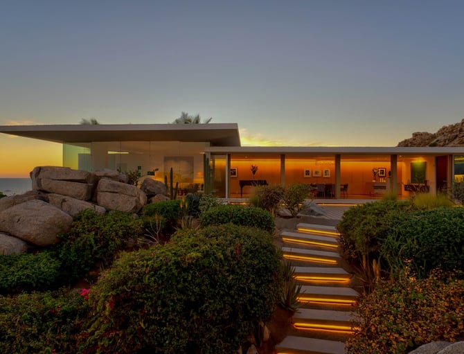 Casa moderna de una planta con paredes de cristal iluminadas al atardecer, rodeada de vegetación desértica, grandes rocas y un sendero iluminado con vistas al mar al ponerse el sol.